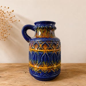 Peut inclure: Un vase en céramique bleu avec une anse, présentant une surface texturée avec des motifs géométriques et en forme de feuilles en orange et jaune. Le vase est posé sur une surface en bois.