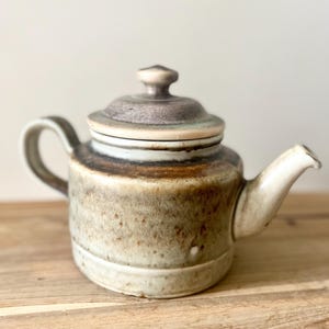 Peut inclure: Une théière en céramique avec un couvercle, une anse et un bec verseur. La théière est principalement beige avec des accents marron et blancs. Le couvercle a un petit bouton sur le dessus. La théière est posée sur une surface en bois.