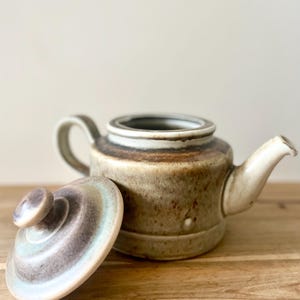 Peut inclure: Une théière en céramique avec un couvercle, présentant un glaçage marron et beige moucheté. Le couvercle est sur le côté, révélant un motif en couches avec des nuances de bleu, de gris et de blanc. La théière a une anse et un bec courbés.