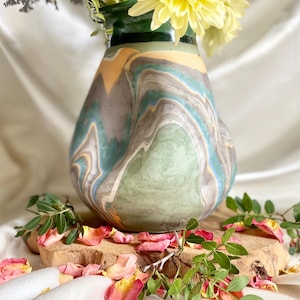 Peut inclure: Un vase décoratif en céramique avec un motif marbré dans les tons de vert, bleu, gris et jaune. Le vase contient des fleurs jaunes. Le vase est posé sur une planche de bois avec des pétales de rose roses et des feuilles vertes.