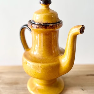 Peut inclure: Une théière en céramique jaune moutarde avec une finition brillante et des accents brun foncé sur la poignée, le bout du bec et le bouton du couvercle. La théière présente une base sur pied, un bec incurvé et un couvercle amovible assorti.