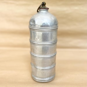 Peut inclure: Une gourde cylindrique en métal argenté présentant une série de rainures horizontales sur le corps. Le sommet de la gourde est muni d'un petit bouchon couleur laiton et d'un anneau métallique circulaire pour la fixation, avec une finition usée.