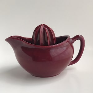 May include: A maroon ceramic citrus juicer with a separate reamer. The juicer has a handle and a rounded base.