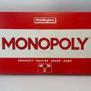 Könnte beinhalten: Eine rot-weiß Monopoly-Brettspielbox. Das Wort "MONOPOLY" steht in großen roten Buchstaben auf weißem Hintergrund. Die Box zeigt auch das Logo "Waddingtons" und den Text "PROPERTY TRADING BOARD GAME".