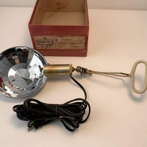 May include: Vintage chrome and brass extension flash unit with a reflector and handle. The unit is connected to a black cord and plug. A red cardboard box with text is in the background.