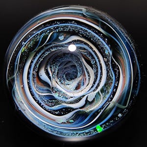 May include: A glass marble with a swirling pattern of blue, white, and black. The marble has a deep, dark center and is covered in iridescent glitter.