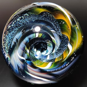 May include: A glass marble with a swirling pattern of blue, green, yellow, and white. The marble has a dark blue background with a white spiral in the center.