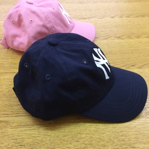 Peut inclure: Deux casquettes de baseball, l'une rose et l'autre bleu marine, toutes deux avec un logo brod&eacute; blanc des New York Yankees.