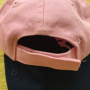 Peut inclure: Une casquette de baseball rose avec une casquette de baseball noire en dessous. La casquette rose a un petit trou sur le dessus et une sangle &agrave; l'arri&egrave;re.