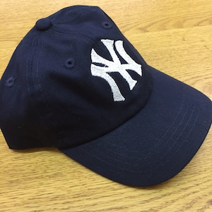 Peut inclure: Une casquette de baseball bleu marine avec un logo brod&eacute; blanc des New York Yankees.
