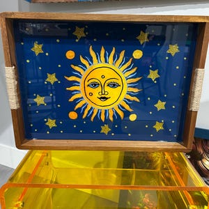 May include: A rectangular wooden tray with a blue background featuring a yellow sun with a smiling face and radiating lines. Gold glitter stars and circles are scattered across the tray. The tray has rope handles.