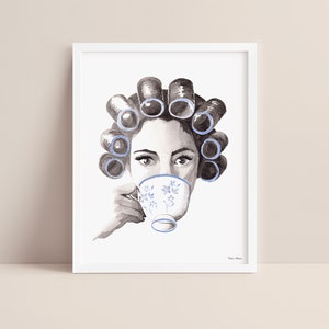 Watercolor portrait of a woman in grayscale with blue curlers in her hair sipping from a blue floral teacup. Print is displayed in a white picture frame on a blush pink background.