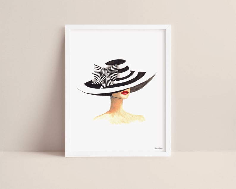 Derby hat art print featuring watercolor portrait of woman wearing black and white striped wide brimmed hat. Print is displayed in a white picture frame on a blush pink background.