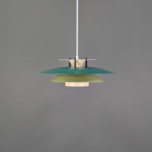 May include: A modern pendant light fixture with a layered design. The light features three circular shades in shades of green and white, suspended from a white cord. The fixture has a minimalist aesthetic, suitable for various interior styles.