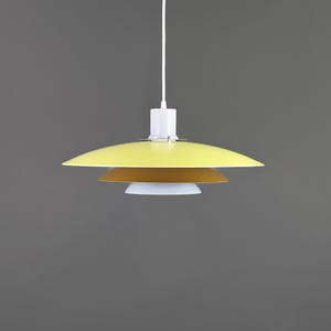 Puede incluir: Una moderna lámpara colgante con un diseño en capas. La pantalla superior es amarilla, seguida de una pantalla marrón y una pantalla blanca en la parte inferior. La lámpara está suspendida de un cable blanco y un soporte de techo blanco.