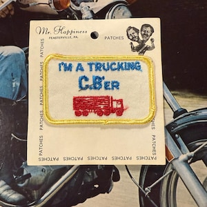May include: A vintage patch with the text "I'M A TRUCKING C.Ber" in blue lettering, and a red truck graphic. The patch is bordered with gold stitching and is on a white background. The patch is attached to a cardboard backing.