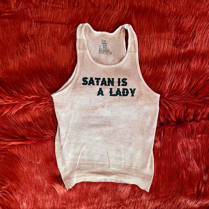 May include: A white tank top with the text "Satan is a Lady" printed in black.