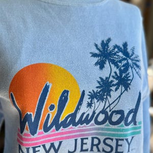 May include: Light blue sweatshirt with a graphic of a sunset, palm trees, and the text "Wildwood NEW JERSEY" in navy blue. The sunset is a gradient of orange and yellow. The text is in a retro font.