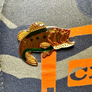 May include: A gold-colored fish pin with black and green accents. The pin features an open mouth and detailed scales. The pin is set against a camouflage pattern.