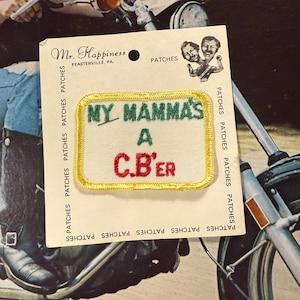 May include: A rectangular embroidered patch with the text "MY MAMMA'S A C.B'ER" in green and red on a white background, with a yellow border. The patch is on a cardboard backing with the "Mr. Happiness" logo and the word "PATCHES".