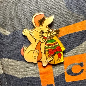 May include: A gold-colored Easter bunny pin holding a decorated egg. The bunny has pink accents on its ears and paws. The egg is green, yellow, and red, with a red bow. The pin is set against a patterned background.