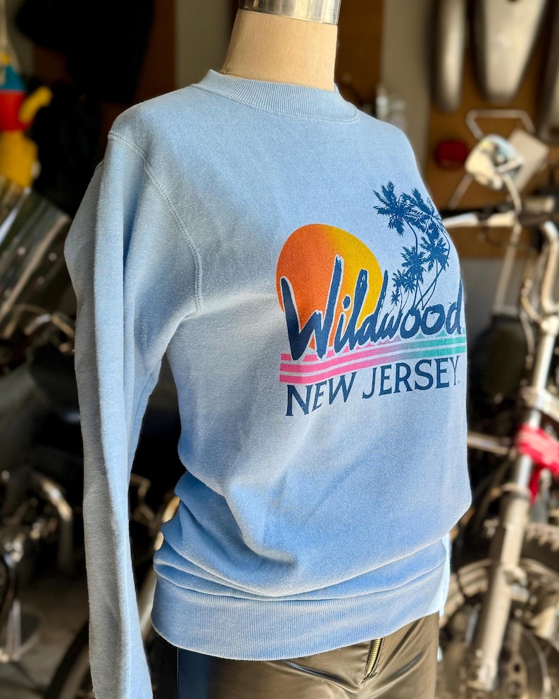 May include: Light blue sweatshirt with the text "Wildwood NEW JERSEY" in a colorful design featuring a sunset and palm trees. The sweatshirt is displayed on a mannequin.