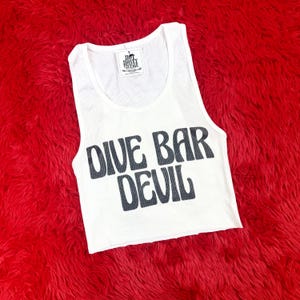 May include: White ribbed tank top with the words "DIVE BAR DEVIL" printed in black. The top has a cropped fit and a tag that reads "HOT HONEY VINTAGE". The tank top is on a red, fuzzy surface.