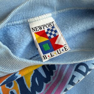 May include: Light blue sweatshirt with a ribbed neckline. The tag reads "NEWPORT BLUE" with a colorful logo. The sleeve features a graphic design with the word "WAVERIDER" in a retro font. The sweatshirt is made of a soft material.