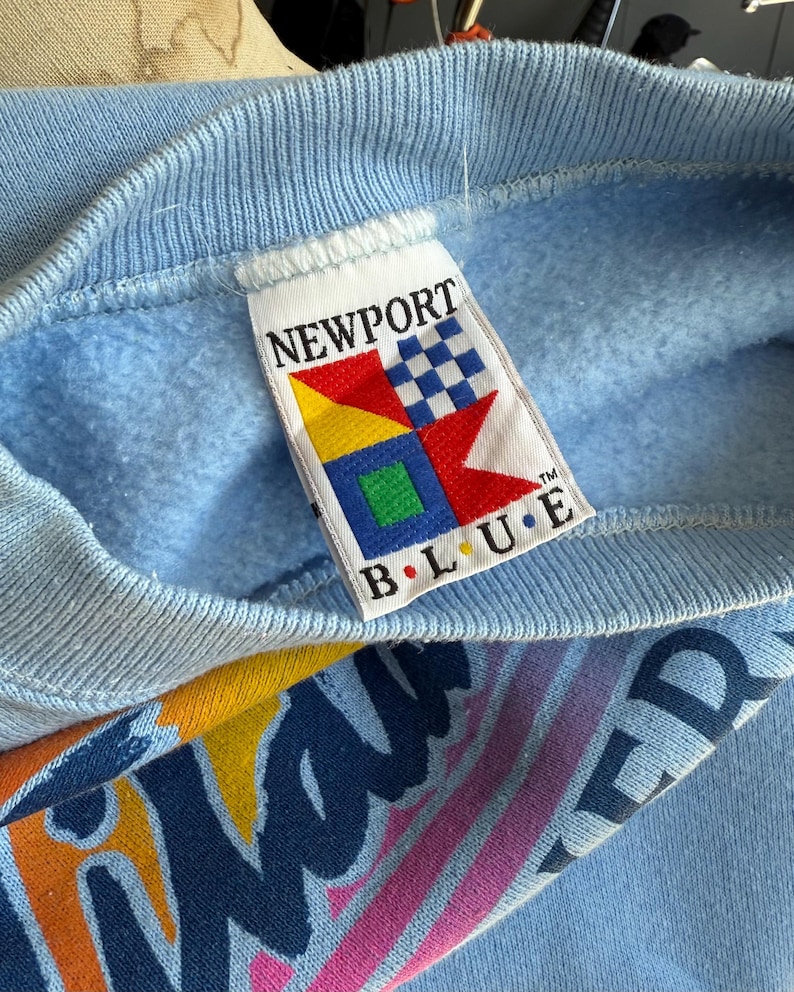 May include: Light blue sweatshirt with a ribbed neckline. The tag reads "NEWPORT BLUE" with a colorful logo. The sleeve features a graphic design with the word "WAVERIDER" in a retro font. The sweatshirt is made of a soft material.