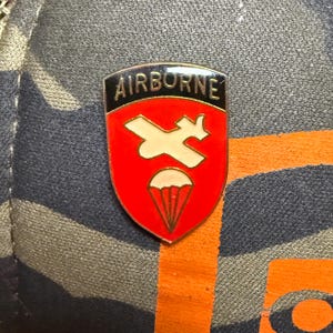 Puede incluir: Un pin de esmalte rojo y blanco con la palabra "AIRBORNE" en la parte superior. El pin presenta un avión blanco y un diseño de paracaídas. El pin está sobre un fondo de camuflaje.