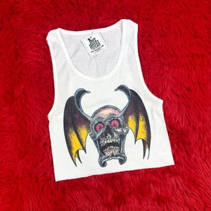May include: White ribbed vest top featuring a skull graphic with bat wings, red eyes, and yellow detailing. The vest has a tag that reads "Hot Honey Vintage".