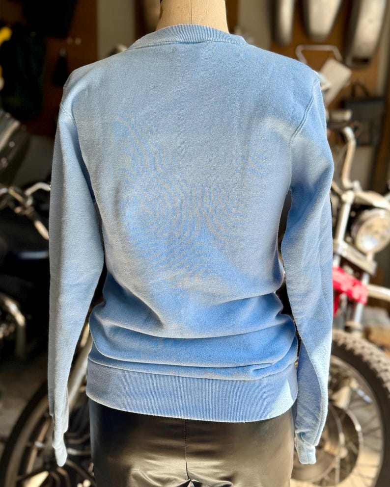 May include: A light blue sweatshirt with a crew neck and long sleeves. The back of the sweatshirt is visible, showing the soft fabric and ribbed hem. The garment is displayed on a mannequin.