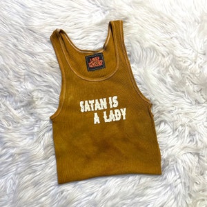 May include: A mustard yellow ribbed tank top with the text "SATAN IS A LADY" printed in white. The tank top has a white tag with the text "HOT HONEY" on it.