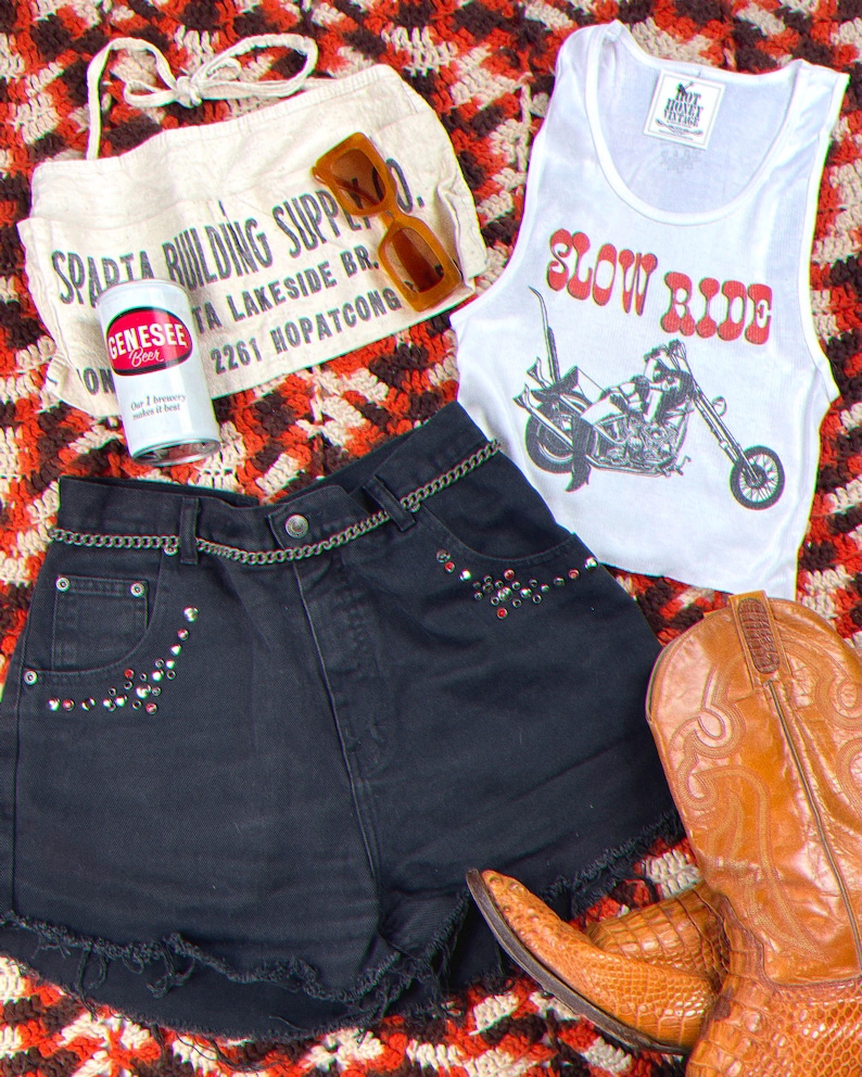 May include: A white tank top with the text "Slow Ride" in red letters and a motorcycle graphic. The tank top is paired with black denim shorts with rhinestones and brown cowboy boots. A white canvas bag with the text "Sparta Building Supply Co." is in the background.