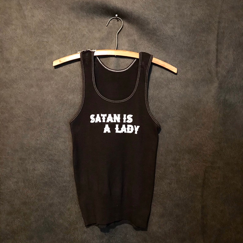 May include: Black tank top with white stitching and the text "SATAN IS A LADY" in white block letters.