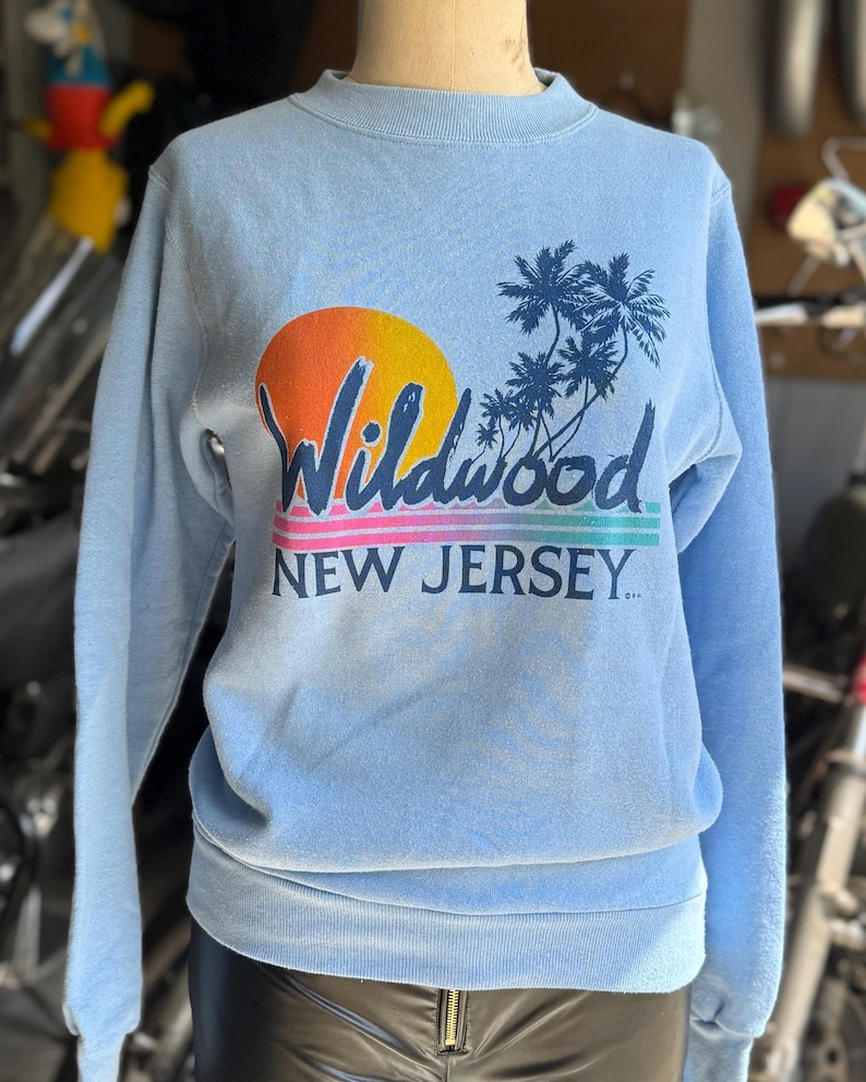 May include: Light blue sweatshirt featuring a sunset graphic with the word "Wildwood" in a script font, palm trees, and the text "NEW JERSEY." The design includes orange, yellow, pink, and blue hues. The sweatshirt has a crew neck and long sleeves.