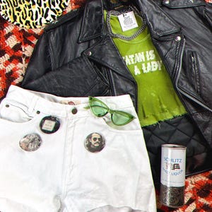 May include: A white denim cut-off shorts with three button pins, a green pair of sunglasses, and a black leather jacket with a green tank top underneath that says "Satan is a Lady".