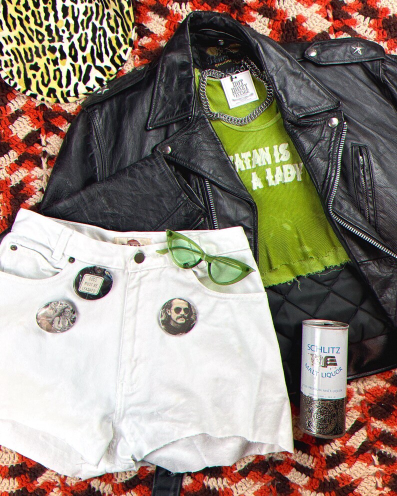 May include: A white denim cut-off shorts with three button pins, a green pair of sunglasses, and a black leather jacket with a green tank top underneath that says "Satan is a Lady".