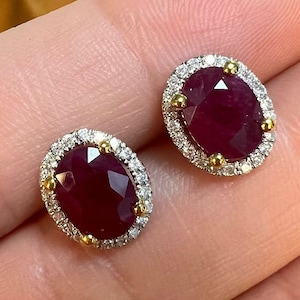 May include: A pair of oval ruby earrings with a halo of small diamonds. The rubies are a deep red color and the diamonds are white. The earrings are set in a white metal with yellow gold accents.