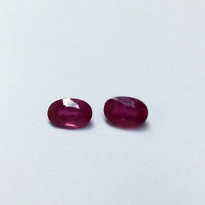 May include: Two oval-cut, deep red rubies are displayed against a white background. The gemstones have a polished, faceted surface, reflecting light. The rubies are of similar size and shape, showcasing their vibrant color and clarity.