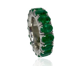 May include: A silver ring featuring a continuous row of vibrant green emeralds. The emeralds are oval-cut and set closely together, creating a full band of color. The ring is displayed against a white background.
