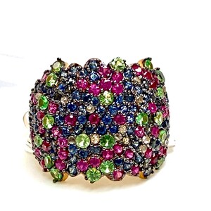 May include: A wide gold band ring with a cluster of colorful gemstones. The ring is set with rubies, sapphires, emeralds, and diamonds. The gemstones are arranged in a random pattern, creating a unique and eye-catching design.