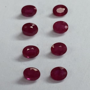 May include: Eight oval-shaped ruby gemstones, each with a deep red color and a faceted cut. The gemstones are arranged in two rows of four, on a white background.