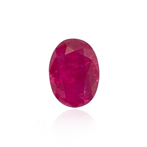 May include: A faceted, oval-shaped ruby gemstone. The gem is a deep, vibrant red color, with a polished surface that reflects light. The ruby is set against a white background, highlighting its color and clarity.