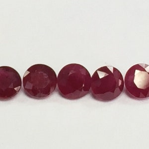 May include: Five round, red gemstones with a faceted cut. The gemstones are arranged in a row on a white surface.
