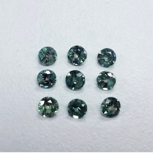 May include: Ten round, faceted gemstones in shades of green and blue. The gemstones are arranged in three rows on a white surface.