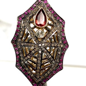May include: A silver ring with a spiderweb design set with pink and white gemstones. The ring is surrounded by a border of pink gemstones.