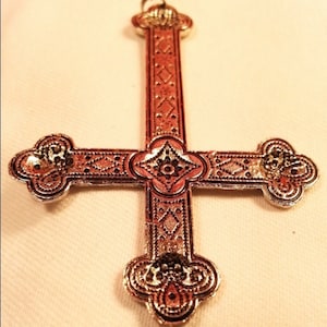 May include: A silver-colored upside-down cross pendant with an intricate, ornate design. The cross has a diamond-shaped pattern on the vertical beam and a floral design on the horizontal beam.