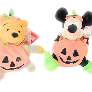 May include: Two plush toys: Winnie the Pooh and Mickey Mouse, both dressed in Halloween pumpkin costumes. The pumpkins are orange with black jack-o'-lantern faces. Pooh has green and pink striped sleeves and legs. Mickey has green feet.