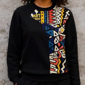 May include: Black sweatshirt with a colorful geometric design on the right side and the words "ART DEPT" in a diamond shape on the left. The design incorporates triangles, zigzags, and other shapes in red, blue, yellow, and white.